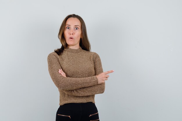 Young woman in golden blouse pointing aside and looking dumbfounded