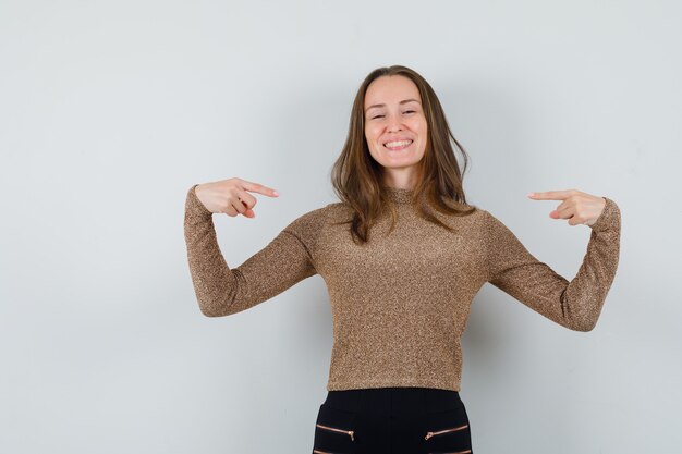 Young woman in golden blouse pointing at herself and looking glad