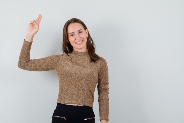 Giovane donna in camicetta dorata che mostra il gesto di addio e che sembra positiva, vista frontale. spazio libero per il testo