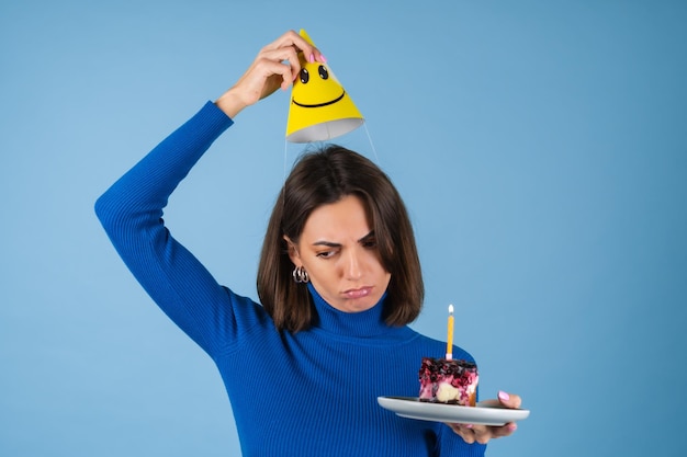 Young woman in a golf on a blue wall celebrates a birthday, holds a piece of cake, is unhappy, disappointed, does not want to grow old