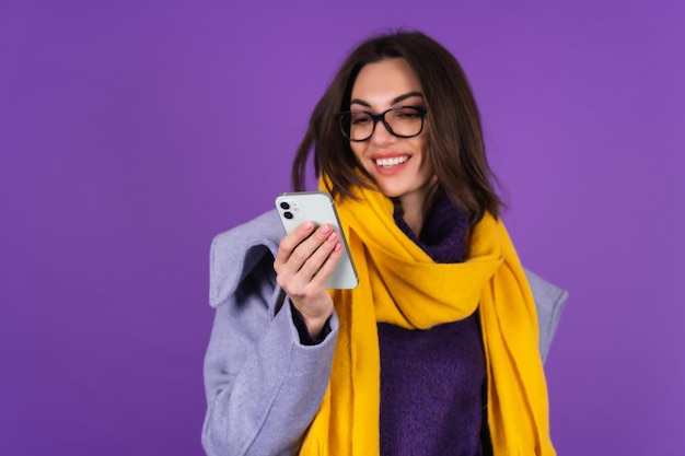 灰色のコート、ニットのドレス、紫色の背景に黄色のスカーフ、ファッショナブルなメガネの若い女性は、電話の画面を見て、元気に笑います