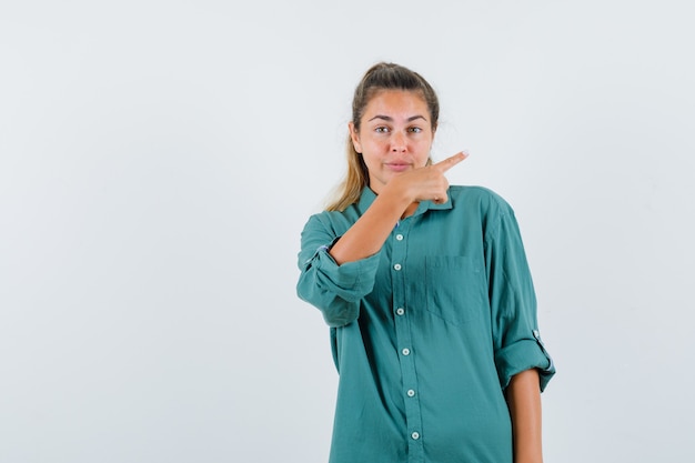 Young woman in green blouse pointing right with index finger and looking happy