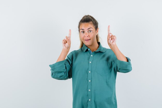 Young woman in green blouse pointing up with index fingers and looking cute