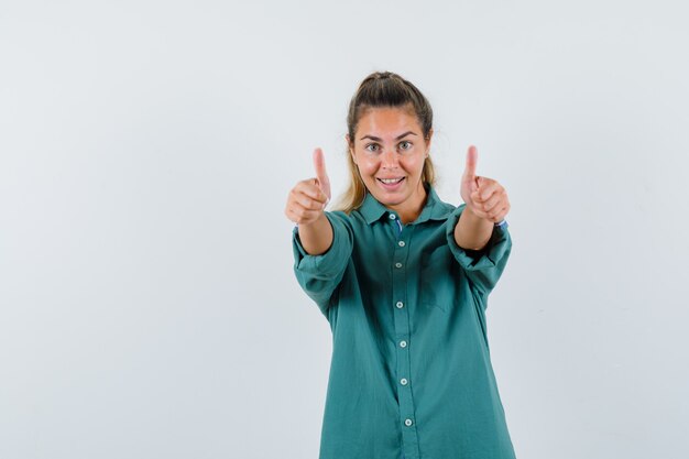 Young woman in green blouse showing thumbs up with both hands and looking happy