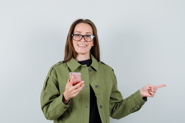 Young woman in green jacket,glasses pointing aside while holding phone , front view.