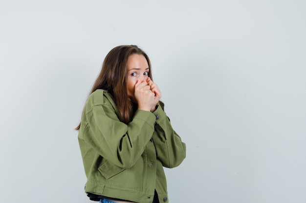 Young woman in green jacket holding fists on her mouth and looking excited , front view.