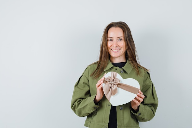 Young woman in green jacket holding heart-shaped box and looking delightful , front view.