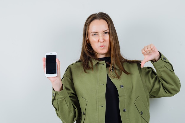 Foto gratuita giovane donna in giacca verde che tiene il telefono mentre mostra il pollice verso il basso e sembra scontento, vista frontale.