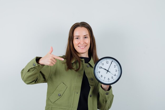 Young woman in green jacket pointing at clock and looking hilarious , front view.