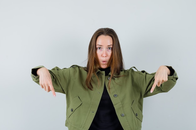 Free photo young woman in green jacket pointing down and looking worried , front view.