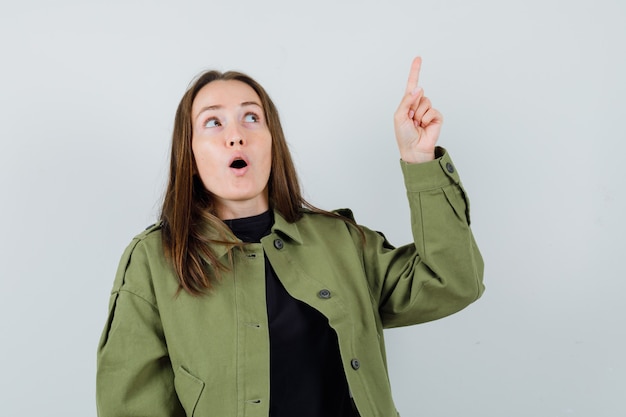 Young woman in green jacket pointing up and looking focused , front view.