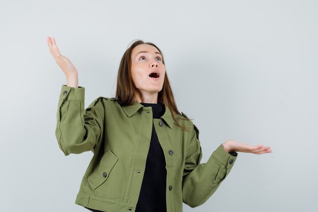 Young woman in green jacket raising hands in questioning manner and looking confused , front view.