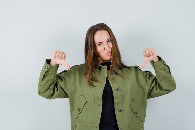 Young woman in green jacket showing thumb down and looking displeased , front view.