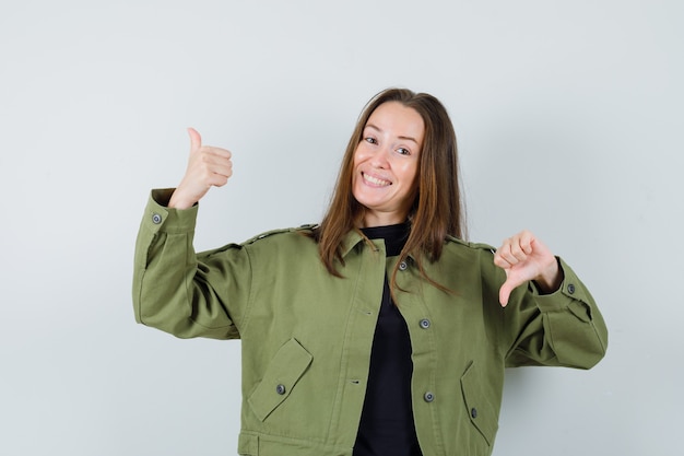 Young woman in green jacket showing thumb up and down and looking joyful , front view.