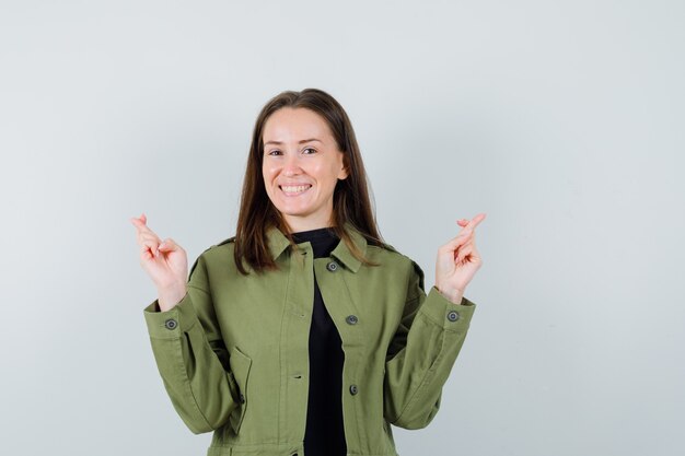 Young woman in green jacket standing with crossed fingers and looking cheery , front view.