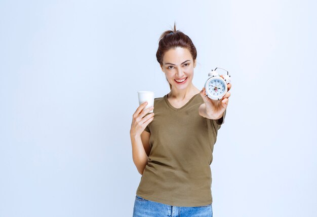 Young woman in green shirt holding an alarm clock and a cup of drink