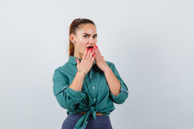 Free photo young woman in green shirt with hands on open mouth and looking surprised , front view.