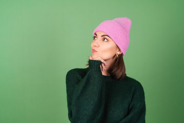 따뜻하고 아늑한 스웨터와 분홍색 모자를 쓴 녹색 옷을 입은 젊은 여성은 귀엽게 웃고, 사려 깊게 옆으로 바라보고, 영감을 받았습니다