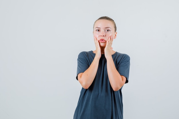 Young woman in grey t-shirt holding hands on cheeks and looking surprised