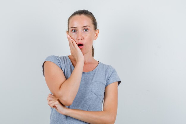 Young woman in grey t-shirt holding palm on cheek and looking shocked , front view.