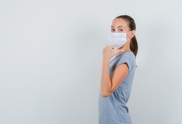 Young woman in grey t-shirt, mask pointing thumb back and looking positive .