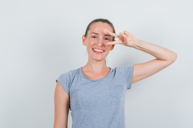 Молодая женщина в серой футболке показывает v-знак возле глаза и радостно смотрит, вид спереди.