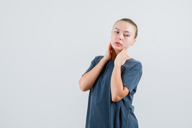 Young woman in grey t-shirt touching neck with hands and looking pensive