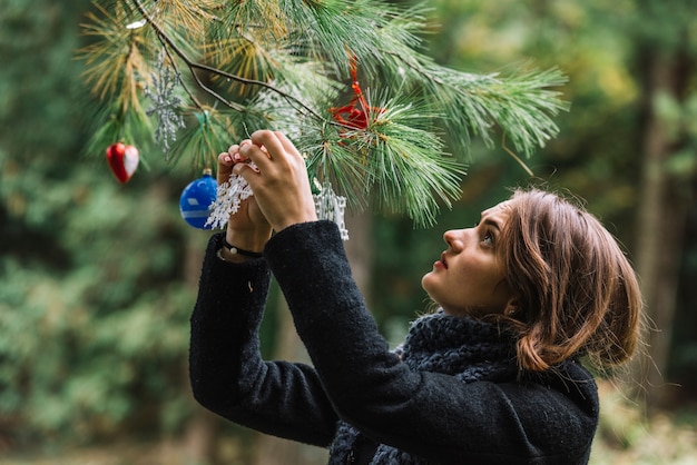 森林の小枝にクリスマスのおもちゃを吊るす若い女性