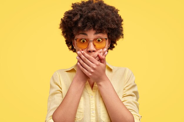 Young woman has scared expression, covers mouth with hands, looks in astonishment, wears trendy shades and blouse