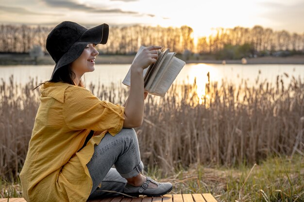 笑顔で帽子をかぶった若い女性が日没時に川のそばに座って本を読んでいます。