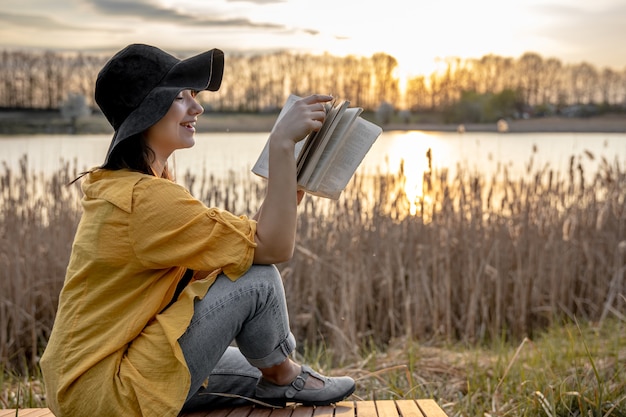 A young woman in a hat with a smile on her face is reading a book sitting by the river at sunset.