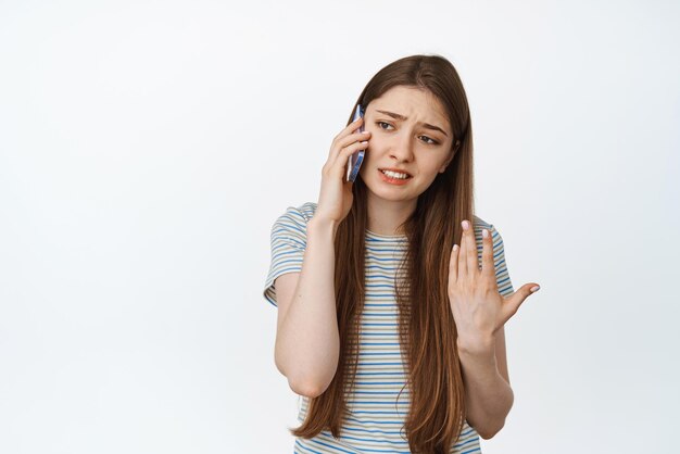 Молодая женщина спорит, объясняя что-то по телефону, разговаривает по мобильному телефону и жестикулирует, стоя на белом фоне