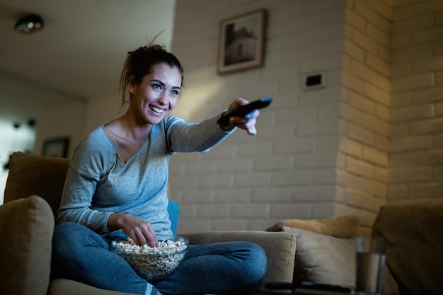 Young woman having fun while changing channels on TV and eating popcorn in the evening at home