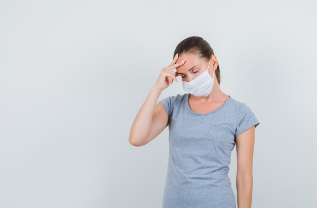 Молодая женщина с головной болью в серой футболке, маске и усталым взглядом, вид спереди.