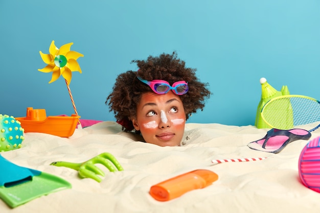 Young woman head with sunscreen cream on face surrounded by beach accessories