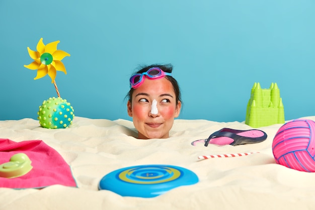 Голова молодой женщины с солнцезащитным кремом на лице в окружении пляжных аксессуаров