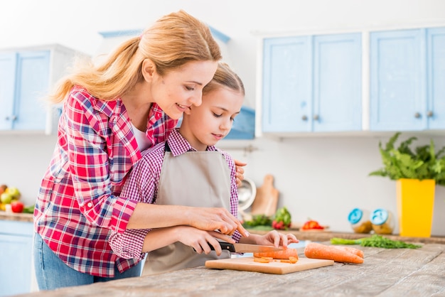 Free photo young woman helping her daughter cutting the carrot with knife on wooden table