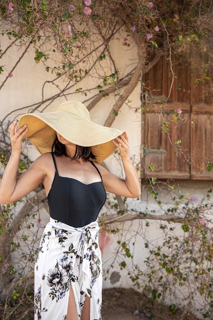 A young woman hides from the sun under a large straw hat near a wall entwined with dry branches of a blossoming tree.