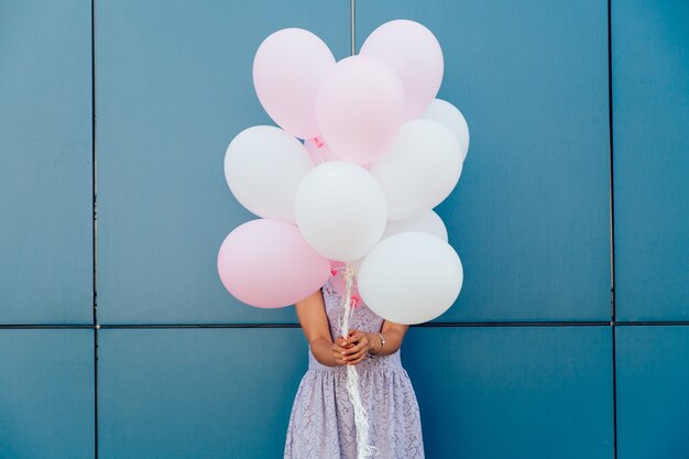 Young woman hiding her face with bunch of balloons, standing against blue wall.