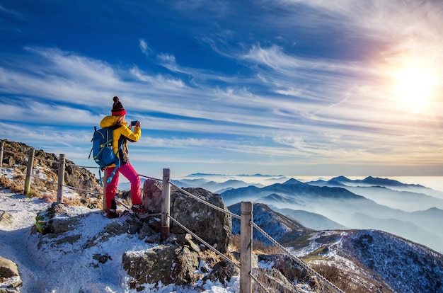 Молодая женщина-путешественница, делающая фото со смартфоном на вершине горы зимой