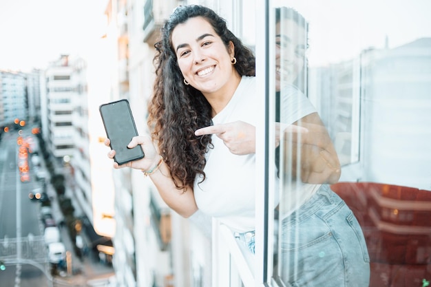 휴대전화를 들고 있는 젊은 여성이 카메라를 가리키며 도시 뒤에 있는 창가를 가리키며 집에서 일하는 젊은 여성 기업가 프리랜서 이미지를 추가할 공간 복사 프리미엄 사진