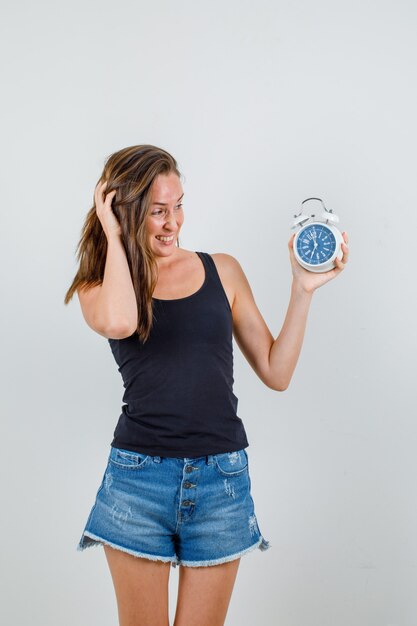 Young woman holding alarm clock in singlet, shorts and looking confused, front view.