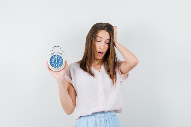 Young woman holding alarm clock in t-shirt, skirt and looking sleepy. front view.