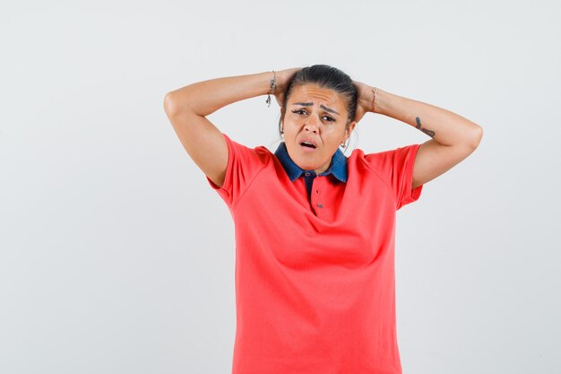 Young woman holding arms behind head in red t-shirt and looking annoyed , front view.