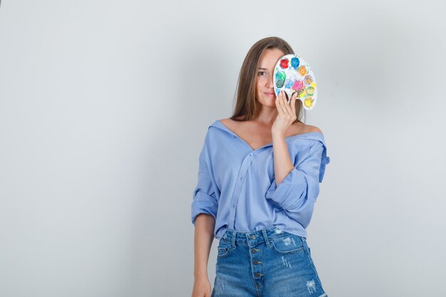 Young woman holding art palette over one eye in blue shirt