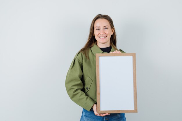 Young woman holding blank frame in green jacket and looking satisfied. front view.