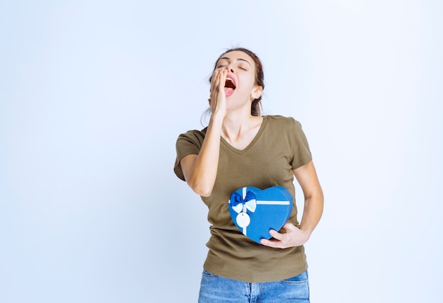 Free photo young woman holding a blue heart shape gift box and shouting out loud