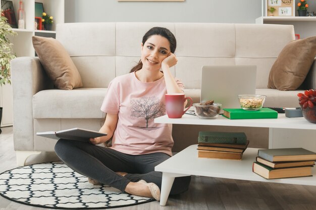 リビングルームのコーヒーテーブルの後ろの床に座って本を保持している若い女性