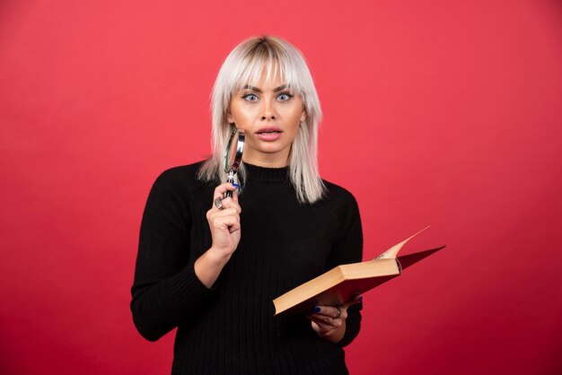 Young woman holding a book with loupe on a red wall. 