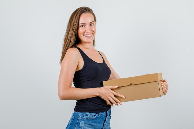 Молодая женщина держит картонную коробку в майке, шортах и выглядит счастливой.